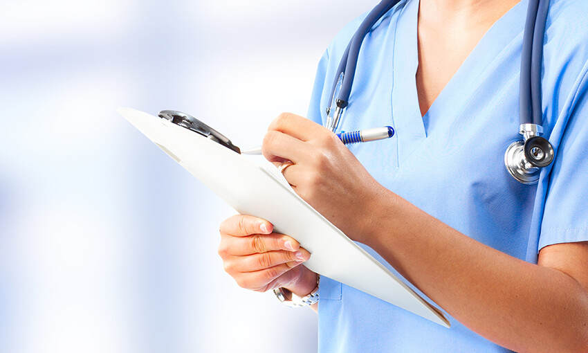 An image of a medical professional wearing blue scrubs holding a clipboard and writing notes.